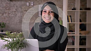 Young beautiful muslim girl in hijab is sitting in office, work concept, waving hello, smile, gesture concept