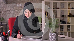 Young beautiful muslim girl in hijab is working with laptop in office, make notes, work concept, business concept