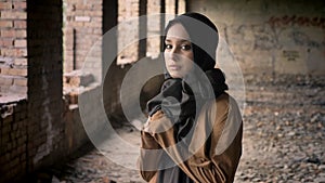 Young beautiful muslim woman in black hijab standing in abandoned building and looking at camera with scared and