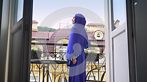Young beautiful muslim woman in hijab going to balcony and standing there, profile of charming lady