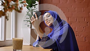 Young beautiful muslim woman in hijab having video chat through smartphone and smiling, sitting in cafe