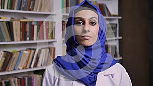Young beautiful muslim woman in hijab standing in library and looking at camera with serious expression