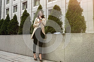 Young beautiful office worker having phone conversation works in her laptop having cup of coffee standing in the street during