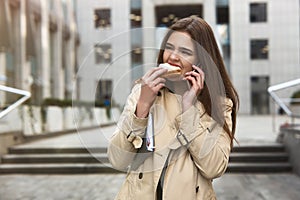 Young beautiful office worker talks on her smartphone walks down the street with planner under her arm bites sandwich in a hurry,