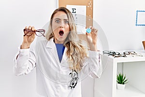 Young beautiful optician woman holding glasses and contact lenses afraid and shocked with surprise and amazed expression, fear and