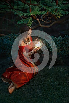 Young beautiful oriental indian fantasy woman sits on green grass, background night magic forest. Red vintage saree