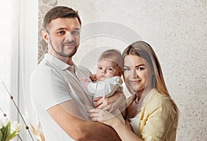 Young beautiful parents hold their newborn daughter in a bright room. A look at the camera