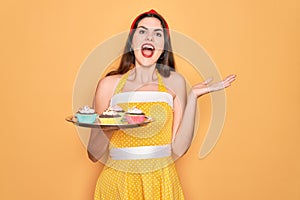 Young beautiful pin up woman wearing 50s fashion vintage dress holding tray with cupcakes very happy and excited, winner