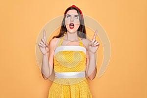 Young beautiful pin up woman wearing 50s fashion vintage dress over yellow background amazed and surprised looking up and pointing