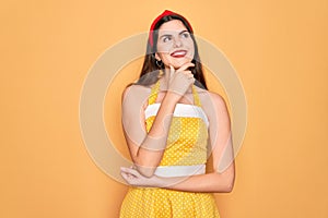 Young beautiful pin up woman wearing 50s fashion vintage dress over yellow background with hand on chin thinking about question,