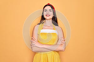 Young beautiful pin up woman wearing 50s fashion vintage dress over yellow background happy face smiling with crossed arms looking