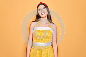 Young beautiful pin up woman wearing 50s fashion vintage dress over yellow background looking away to side with smile on face,