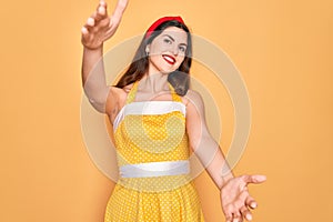 Young beautiful pin up woman wearing 50s fashion vintage dress over yellow background looking at the camera smiling with open arms