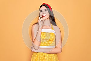 Young beautiful pin up woman wearing 50s fashion vintage dress over yellow background looking confident at the camera with smile