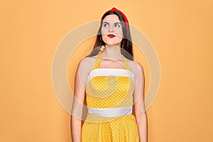 Young beautiful pin up woman wearing 50s fashion vintage dress over yellow background smiling looking to the side and staring away