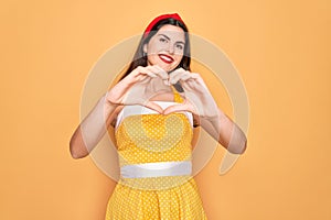 Young beautiful pin up woman wearing 50s fashion vintage dress over yellow background smiling in love doing heart symbol shape