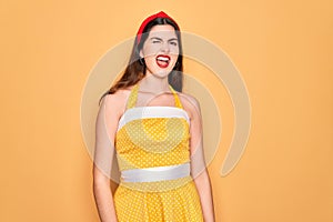 Young beautiful pin up woman wearing 50s fashion vintage dress over yellow background winking looking at the camera with sexy