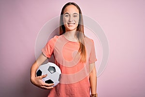 Young beautiful player woman playing soccer holding football ball  over pink background with a happy face standing and smiling