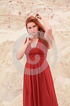 Young beautiful plus size Caucasian woman in red long dress posing in desert landscape with sand.