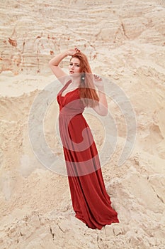 Young beautiful plus size Caucasian woman in red long dress posing in desert landscape with sand.