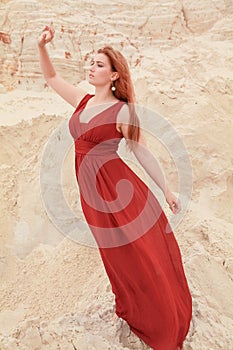 Young beautiful plus size Caucasian woman in red long dress posing in desert landscape with sand.