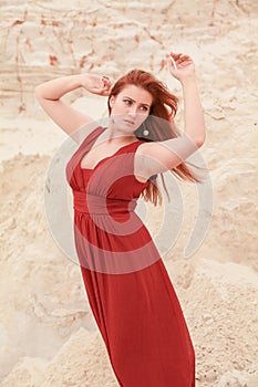 Young beautiful plus size Caucasian woman in red long dress posing in desert landscape with sand.