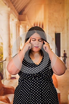 Young beautiful plus size model in black dres having a headache, touching her head bith both hands, xxl woman on gray