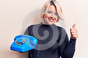 Young beautiful plus size woman holding vintage telephone over isolated white background smiling happy and positive, thumb up