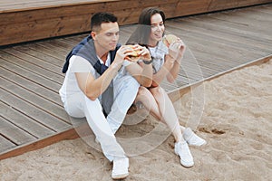 Young beautiful positive couple eating hambergers sitting on beach