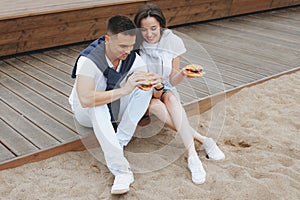 Young beautiful positive couple eating hambergers sitting on beach