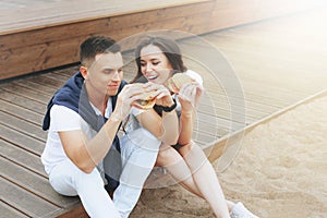 Young beautiful positive couple eating hambergers sitting on beach