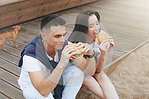 Young beautiful positive couple eating hambergers sitting on beach