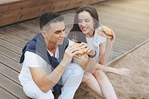 Young beautiful positive couple eating hambergers sitting on beach