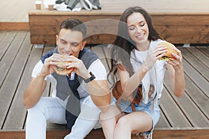 Young beautiful positive couple eating hambergers sitting on beach