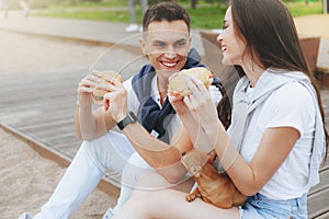 Young beautiful positive couple eating hambergers sitting on beach