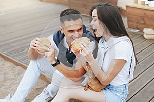 Young beautiful positive couple eating hambergers sitting on beach