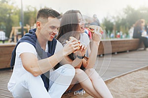 Young beautiful positive couple eating hambergers sitting on beach