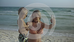 Young beautiful positive mother is sitting with her little son and making a selfie on a smartphone on the beach 4k