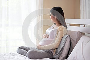Young beautiful pregnant asian woman sitting on bed and touching belly near the window in bedroom. Pregnancy, motherhood, people