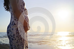 Young beautiful pregnant woman on the beach touching her belly with love and care making a heart shape