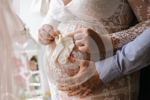 Young beautiful pregnant woman and her husband with hands on her belly keeps white baby socks on her belly.