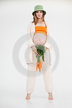 Young beautiful pregnant woman with long dark hair wearing green bucket hat standing, holding fresh bunch of carrots.