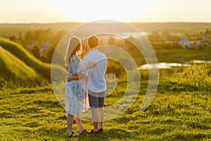 A young beautiful pregnant woman in a long dress and her husband standing together in the hill at sunset