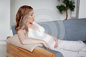 Young Beautiful Pregnant woman sitting on couch at home. Model in white holds hands on belly. Maternity, Expectation, Parenthood