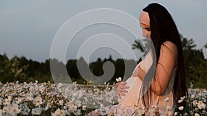 Young beautiful pregnant woman sitting in a flower field stroking her belly and smiling at sunset.