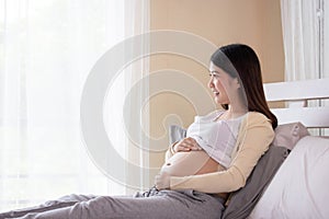 Young beautiful pregnant woman touching belly near the window in bedroom. Pregnancy, motherhood, people and expectation concept