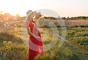 Young beautiful pregnant woman wearing red dress and heat walking in the green field on a sunny summer day