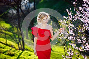 Young beautiful pretty woman posing in long evening luxury dress against bushes with blossoming spring tree. Vogue style