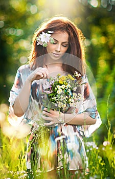 Young beautiful red hair woman holding a wild flowers bouquet in a sunny day. Portrait of attractive long hair female with flowers