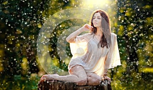 Young beautiful red hair woman wearing a transparent white blouse posing on a stump in a green forest. Fashionable girl